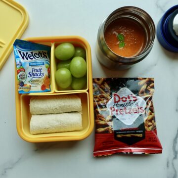 Tomato soup lunchbox with turkey and cheese rollups, grapes, fruit snacks, and pretzels.