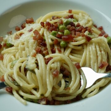 Creamy pasta with pancetta and peas made without cream, topped with crispy pancetta, grated parmesan, and a squeeze of lemon in a white bowl.