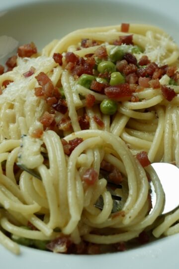 Creamy pasta with pancetta and peas made without cream, topped with crispy pancetta, grated parmesan, and a squeeze of lemon in a white bowl.