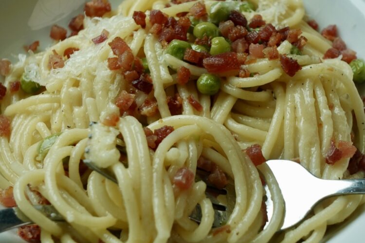 Creamy pasta with pancetta and peas made without cream, topped with crispy pancetta, grated parmesan, and a squeeze of lemon in a white bowl.