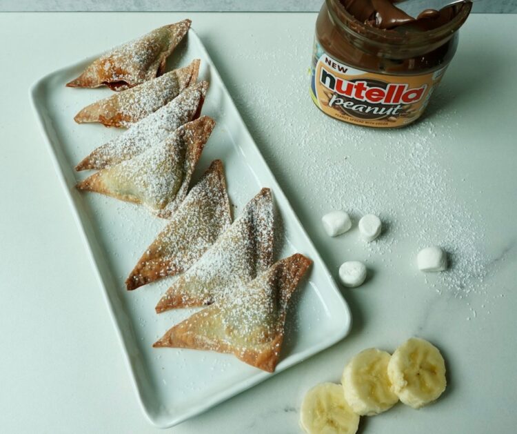 Nutella chocolate dumplings dusted with powdered sugar served on a white plate with banana slices and marshmallows.