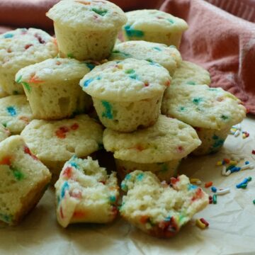 Homemade party cake mini muffins filled with rainbow sprinkles, with one muffin split open to show its soft, moist crumb.