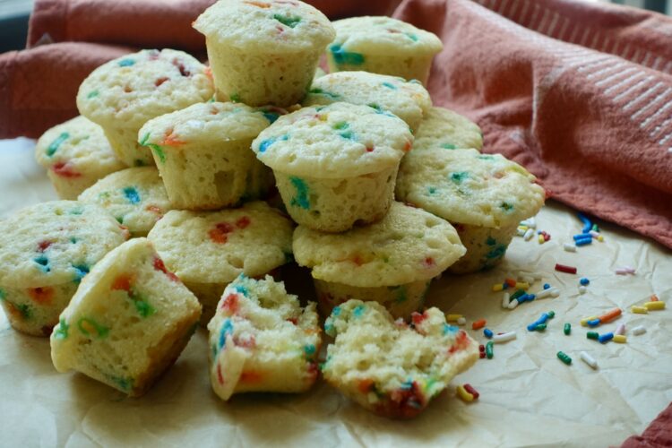 Homemade party cake mini muffins filled with rainbow sprinkles, with one muffin split open to show its soft, moist crumb.